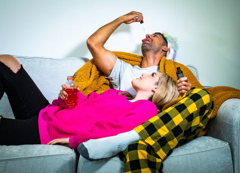 A hungover couple lounges on the couch while the man drops CBD oil in his mouth
