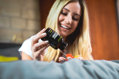 A blond woman pours out some CBD gummies from a bottle of Sunday Scaries while laying on her bed