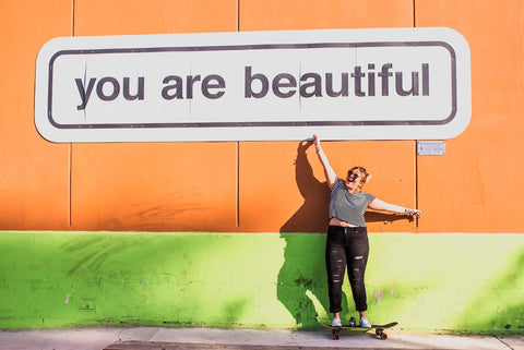 A woman on a skateboard points to a sign on a wall that reads "you are beautiful"