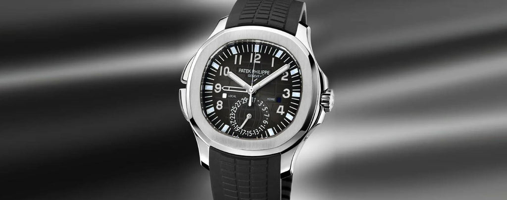 Close-up photo of a Patek Philippe Aquanaut Travel Time watch on a wrist. The watch has a stainless steel case, a black dial with additional subdials for the travel time function, and a black rubber strap.