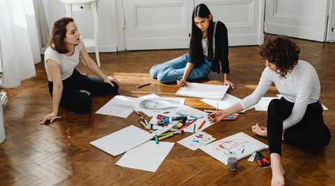 group of girls sitting on the floor all have art supplies out, they are conversating while drawing faces ad designs