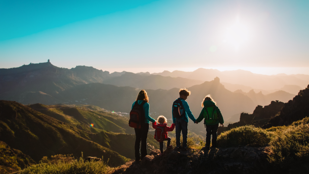 Mom and her three kids wearing puffer jackets on top of the mountain at sunrise while looking down at the view of the town
