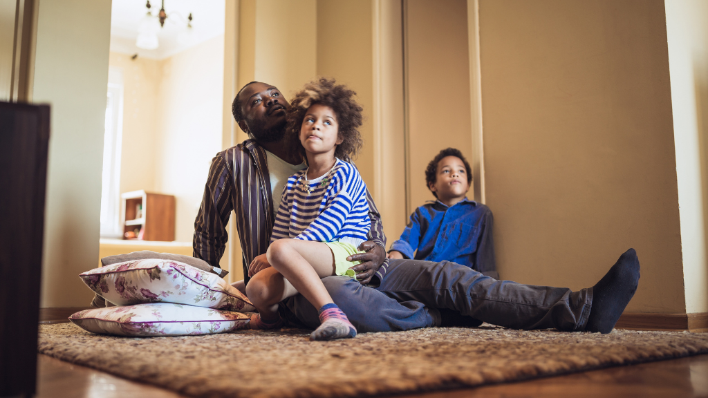 Family sitting on the floor while watching tv. The girl is sitting on her father's lap while the boy is sitting on the floor and leaning on the wall