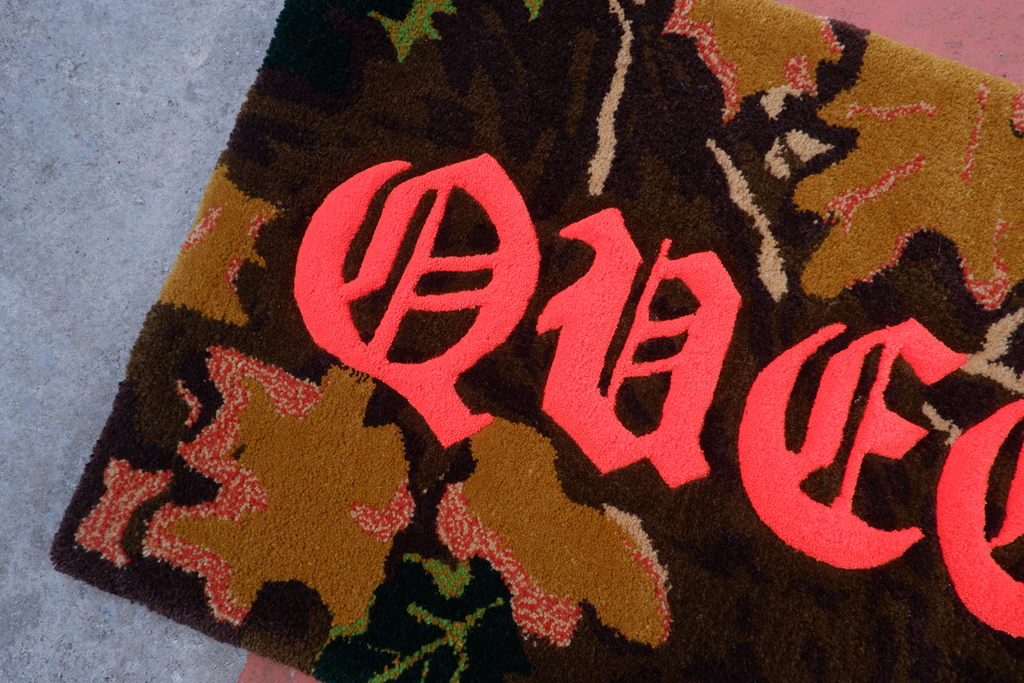 hand-tufted queer rug with neon orange text that says "QUEER" with a camo, RealTree background