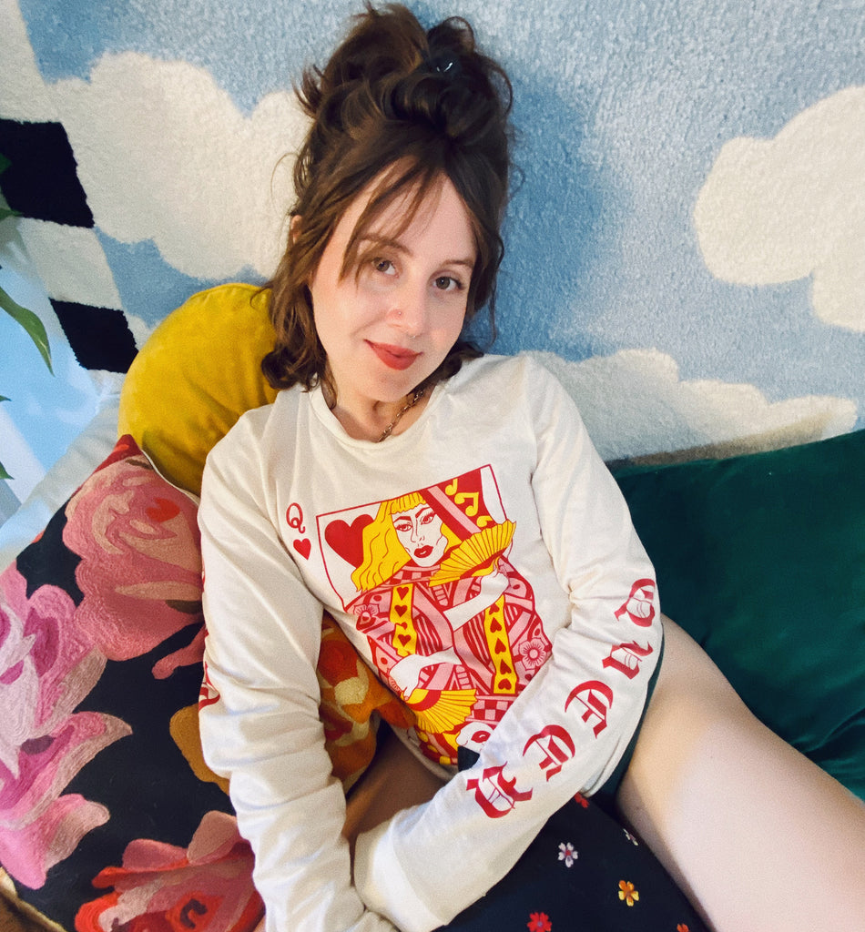 AJ sitting on a bed in front of a rug with sky and clouds wearing the Queer of Hearts long sleeve shirt.