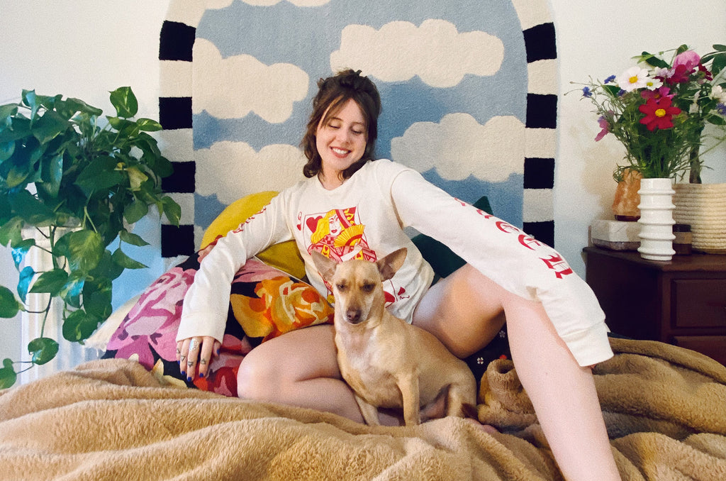 AJ sitting on a bed in front of a rug with sky and clouds wearing the Queer of Hearts long sleeve shirt.