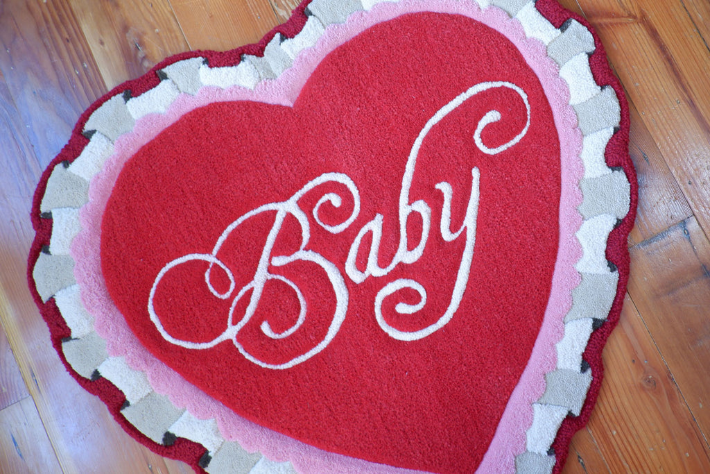 a valentine themed, heart-shaped rug with ruffle detailing. The rug is red and pink and has the word "Baby" in a script font.