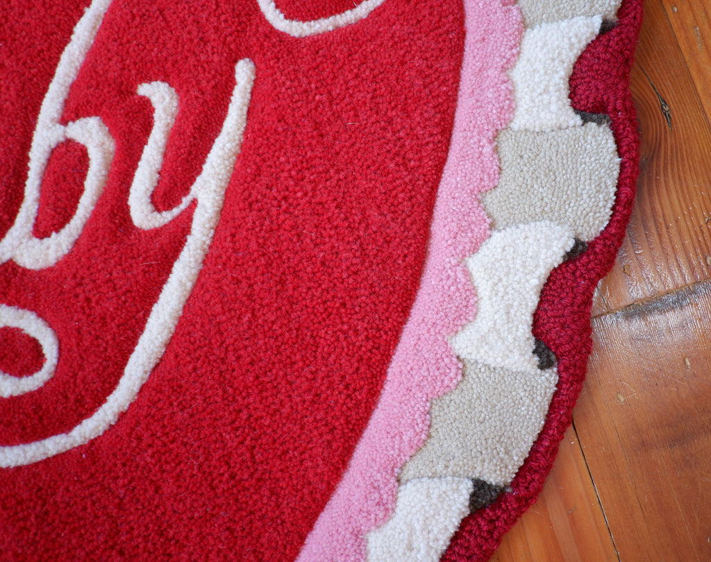 close up of a valentine themed, heart-shaped rug with ruffle detailing. The rug is red and pink and has the word "Baby" in a script font.