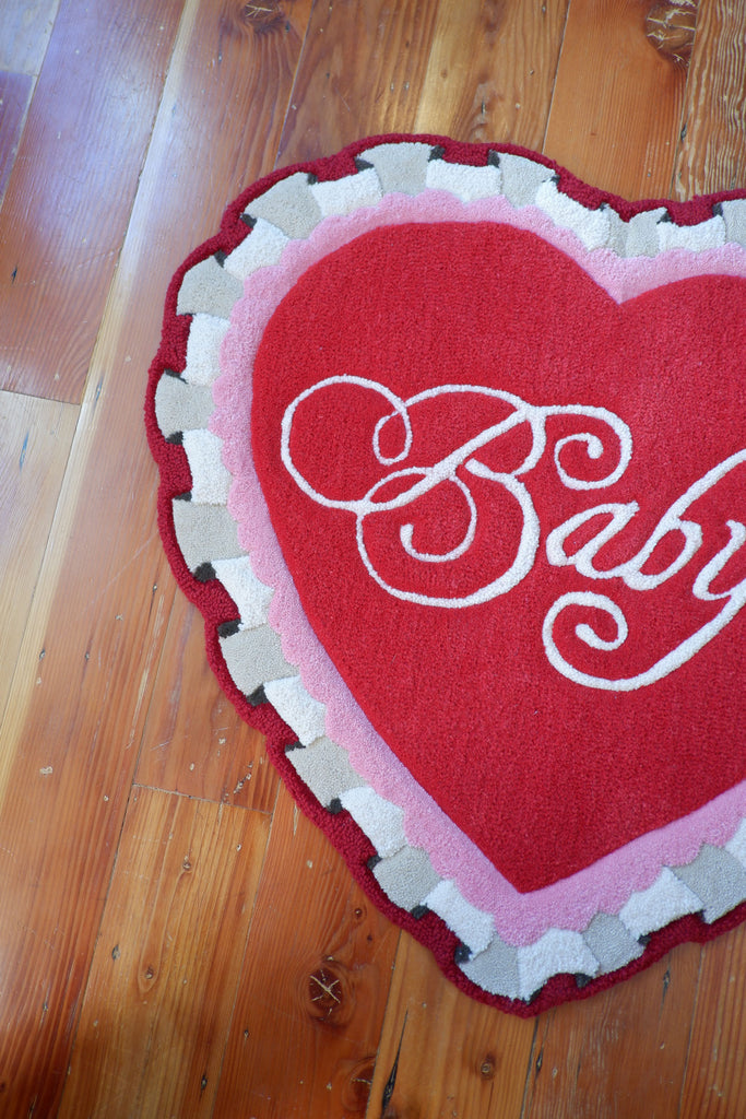 a valentine themed, heart-shaped rug with ruffle detailing. The rug is red and pink and has the word "Baby" in a script font.