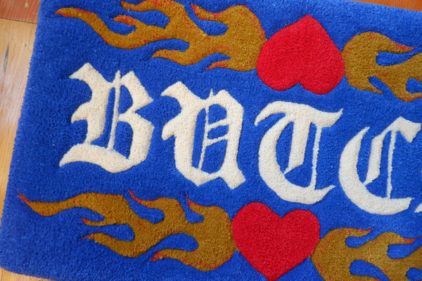 a royal blue hand-tufted queer rug with the word "Butch" on it surrounded by flaming hearts.