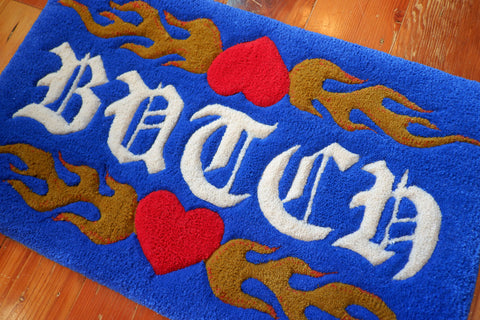 a royal blue hand-tufted queer rug with the word "Femme" on it surrounded by flaming hearts.