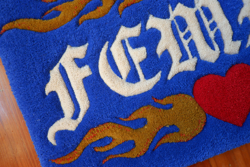 a royal blue hand-tufted queer rug with the word "Femme" on it surrounded by flaming hearts.