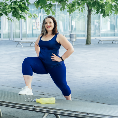 A cheerful person wearing a coordinated blue outfit is posed confidently with one hand on her hip and the other on her thigh, performing a lunge exercise outdoors. A resistance band lies on the bench beside her, suggesting a versatile, low-impact workout routine conducive to spinal health and back pain management