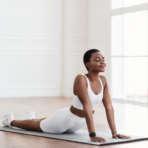 An individual in a white tank top and leggings is performing the Cobra Pose, a yoga stretch beneficial for back health. They are lying face-down on a yoga mat with their upper body gently arched back and elevated by straight arms, allowing for a stretch in the abdominal muscles and a decompression of the spine. The closed eyes and serene expression suggest a focus on deep breathing and relaxation.