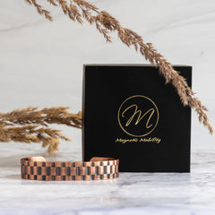 Elegant copper bracelet from Magnetic Mobility with a distinct textured design, displayed in front of a black branded box, accented by dried golden grass on a marble backdrop