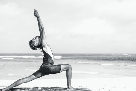 A person in a warrior pose on a beach, with one leg bent at the knee and the other extended back, arms reaching towards the sky in a graceful arch. This pose strengthens core and lower body muscles, promoting back health as part of a holistic wellness routine.