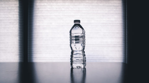A bottle of water is centred on a table, backlit by natural light filtering through a window with blinds, creating a soft and serene ambiance. The bottle casts a crisp shadow on the surface, highlighting the importance of hydration as an essential part of back health maintenance and overall well-being.