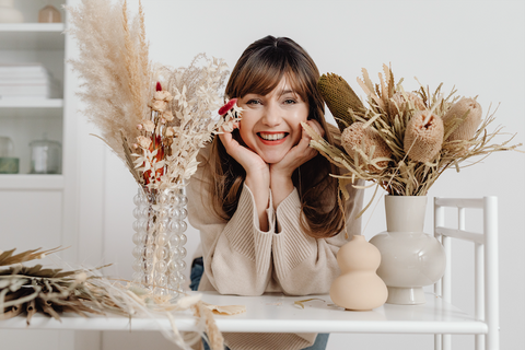 How a Gratitude Journal Can Improve Your Well-being: A woman props her head up on her hands and smiles while surrounded by dried bouquets of flowers.