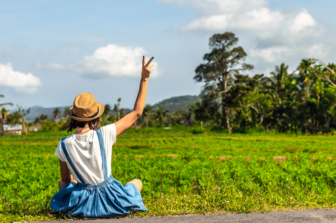 How a Gratitude Journal Can Improve Your Well-being: A woman holds up a peace sign while sitting in front of a field.