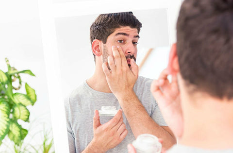 men is applying face cream on his cheeks by looking into the mirror