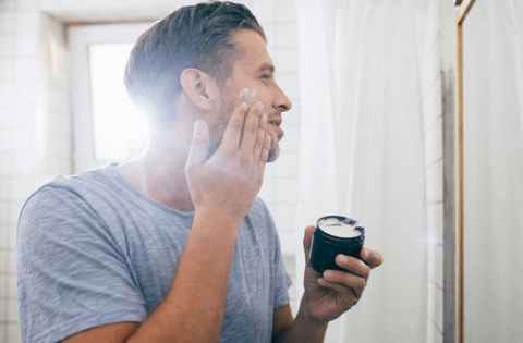 Men is applying face cream with his hands on his face infront of mirror