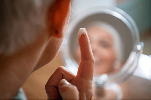 lady is applying anti ageing cream on her cheeks while seeing herself infront of the mirror