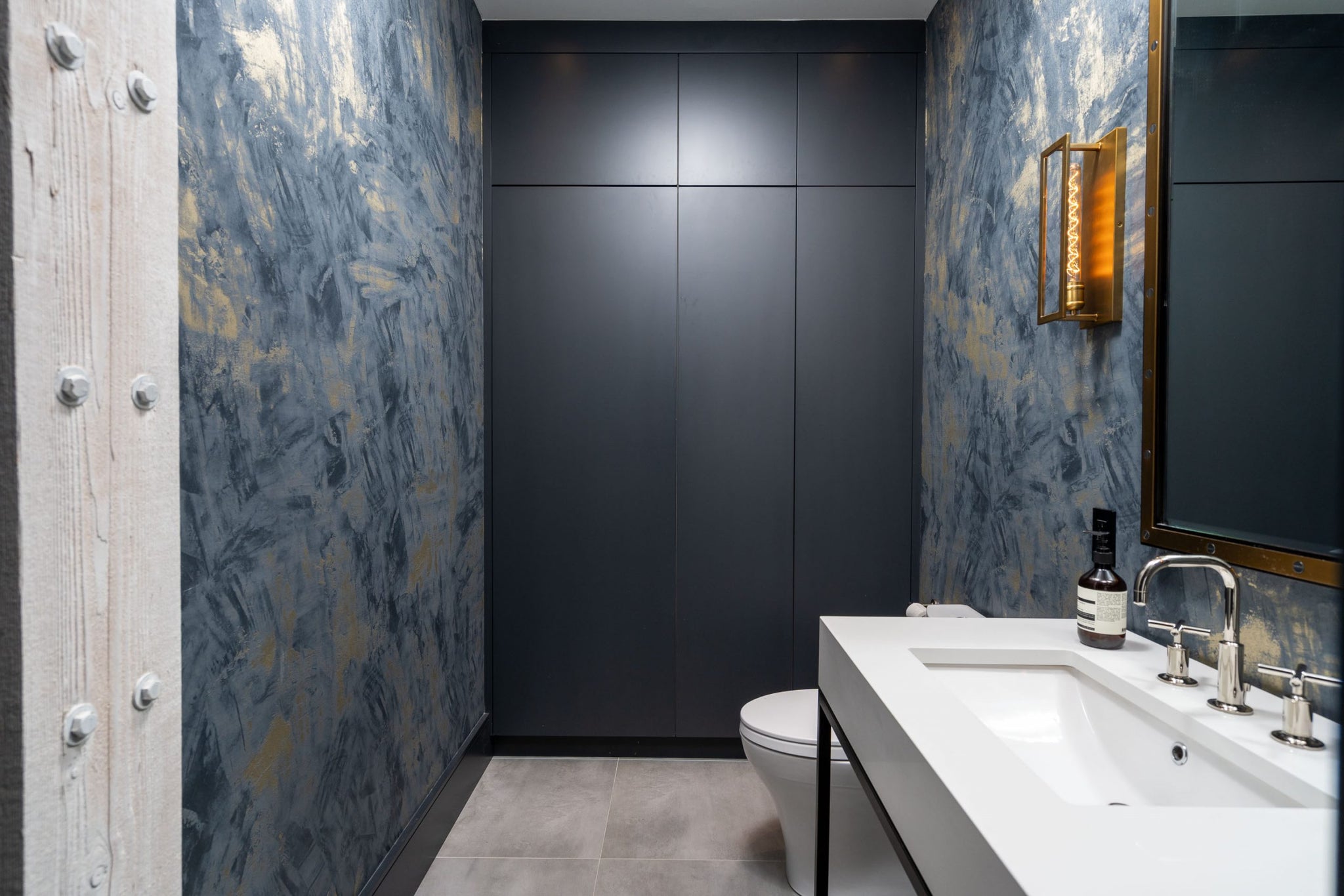 industrial style bathroom design with black framed vanity, rustic door frame, and brass mirror and incandescent vanity lighting