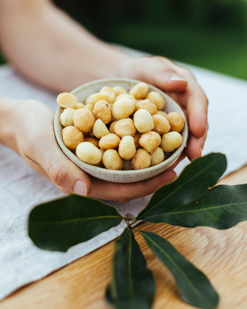 Shelled Macadamia nuts in small bowl held by mans hands. Macadamia leaf below the bowl on a table sitting in the macadamia orchard of the brook family farm Brookfarm