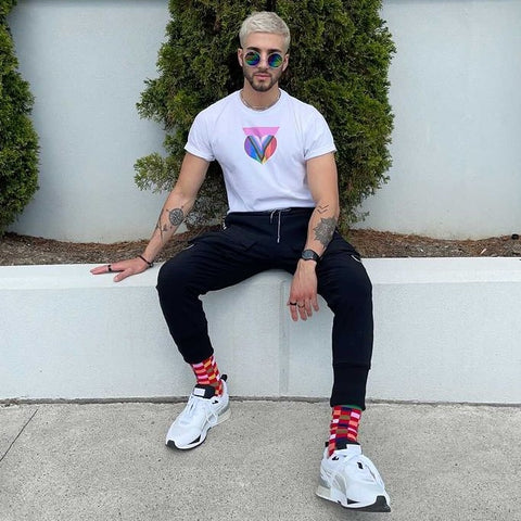 Joe sits in a white Pink Progress Pride Heart design t-shirt with rainbow glasses and colourful socks
