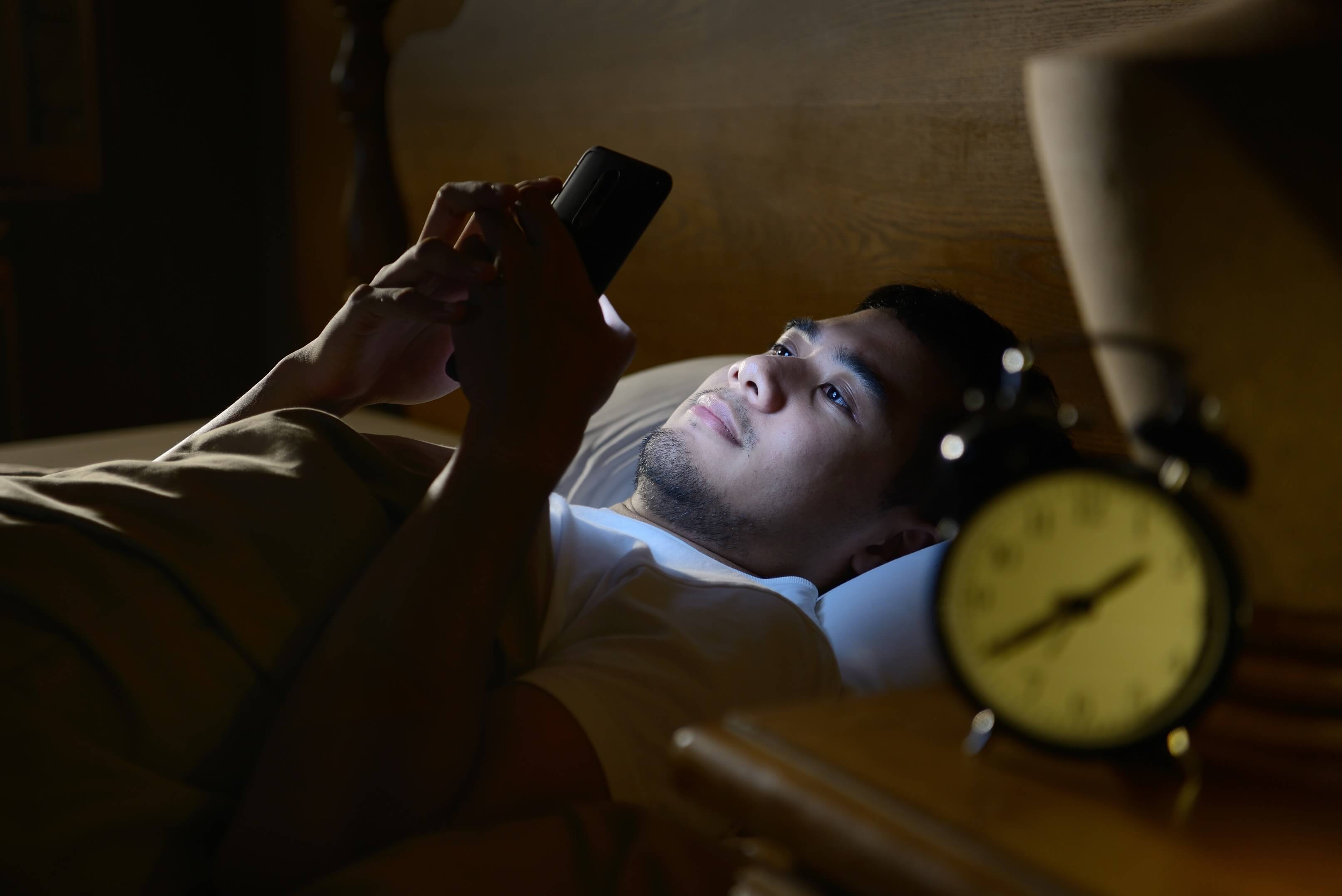 Man laying in bed unable to fall asleep and looking at his brightly lit smartphone.