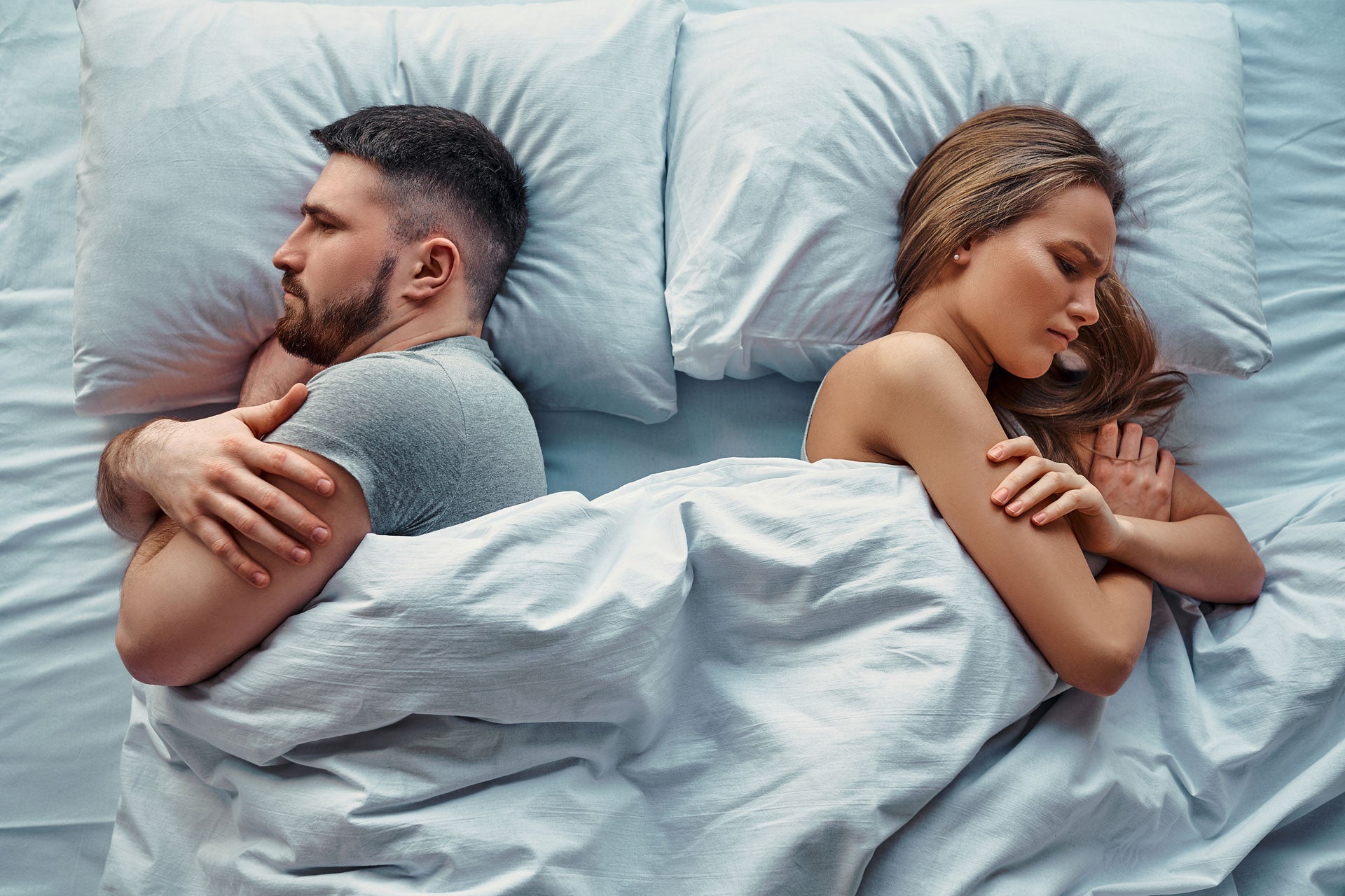 Man and woman sleeping beside each other, facing away from one another looking stressed.