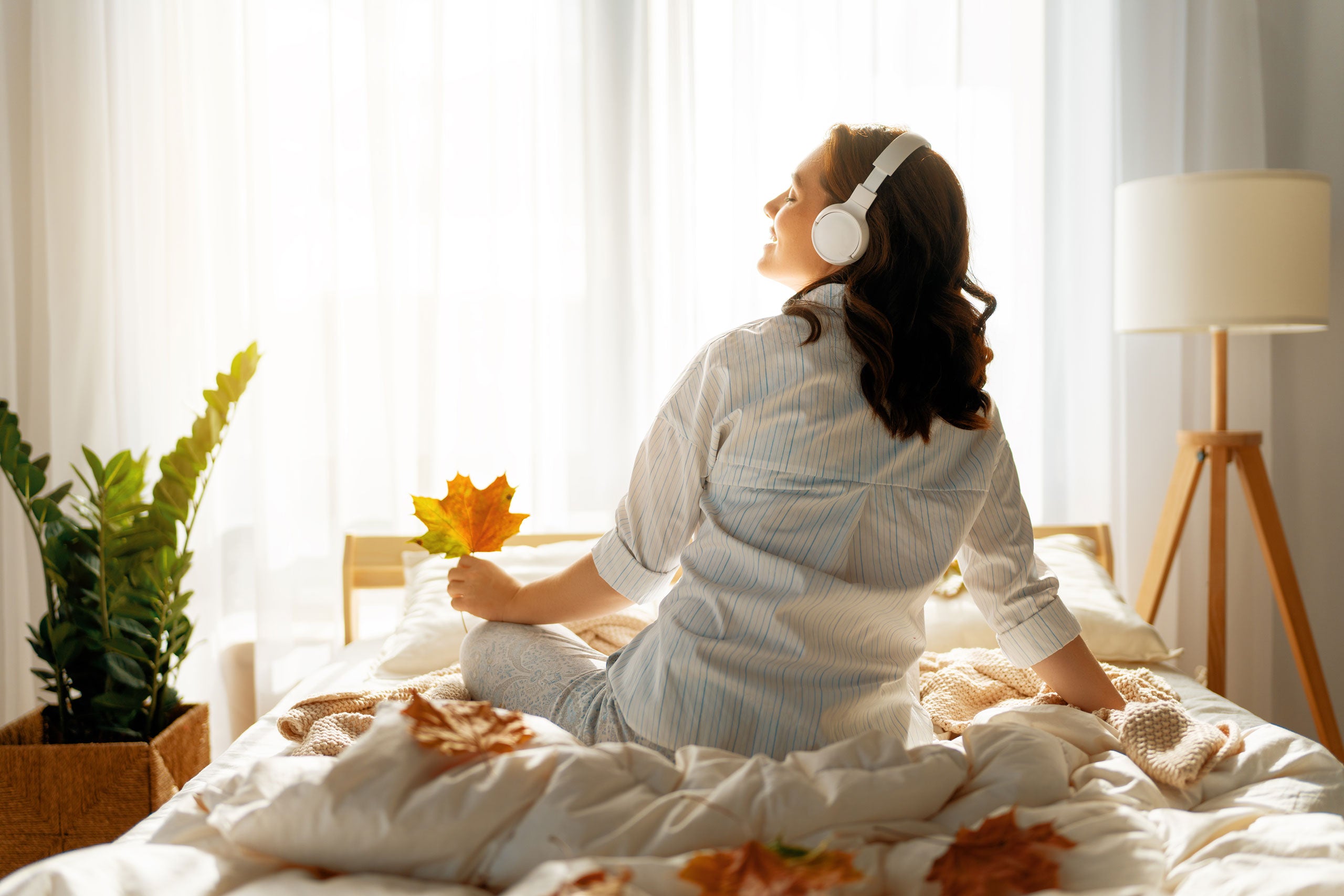 Woman sitting up in bed holding a coloured maple leaf while listening to over-ear headphones.