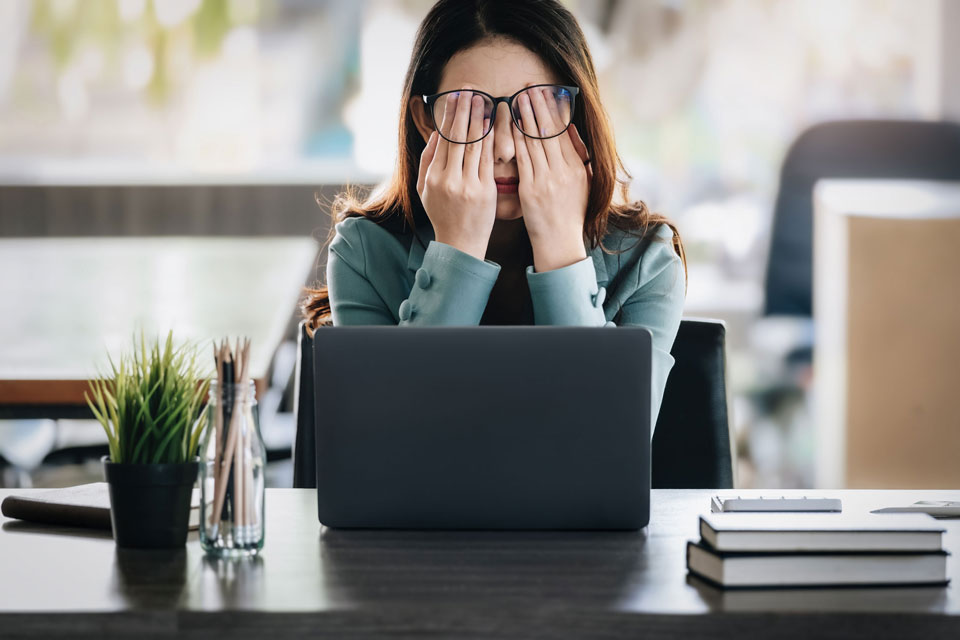 Woman sitting at desk in front of laptop with her hands over her eyes looking tired.