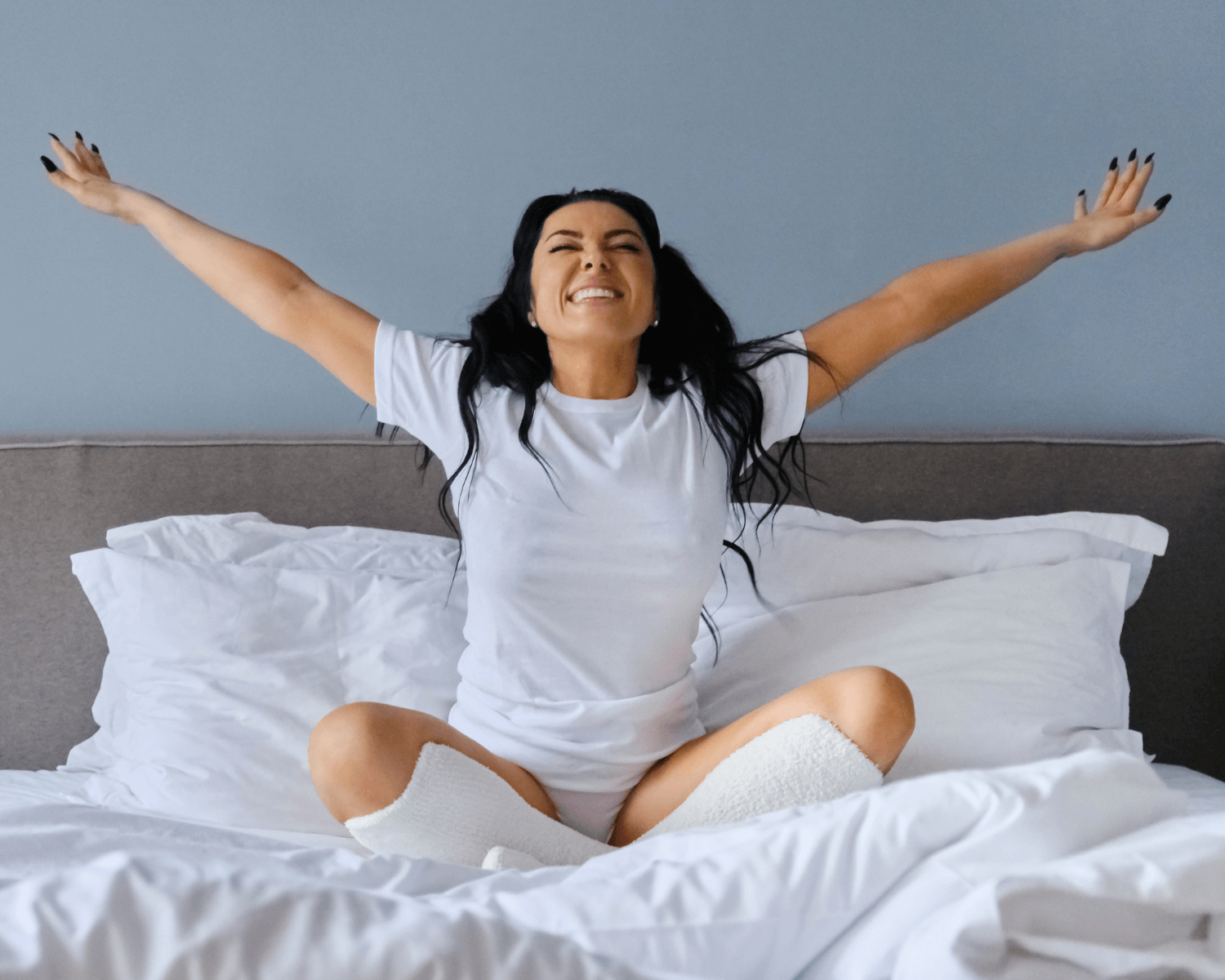 Woman wearing all white stretching her arms out in bed looking happy.