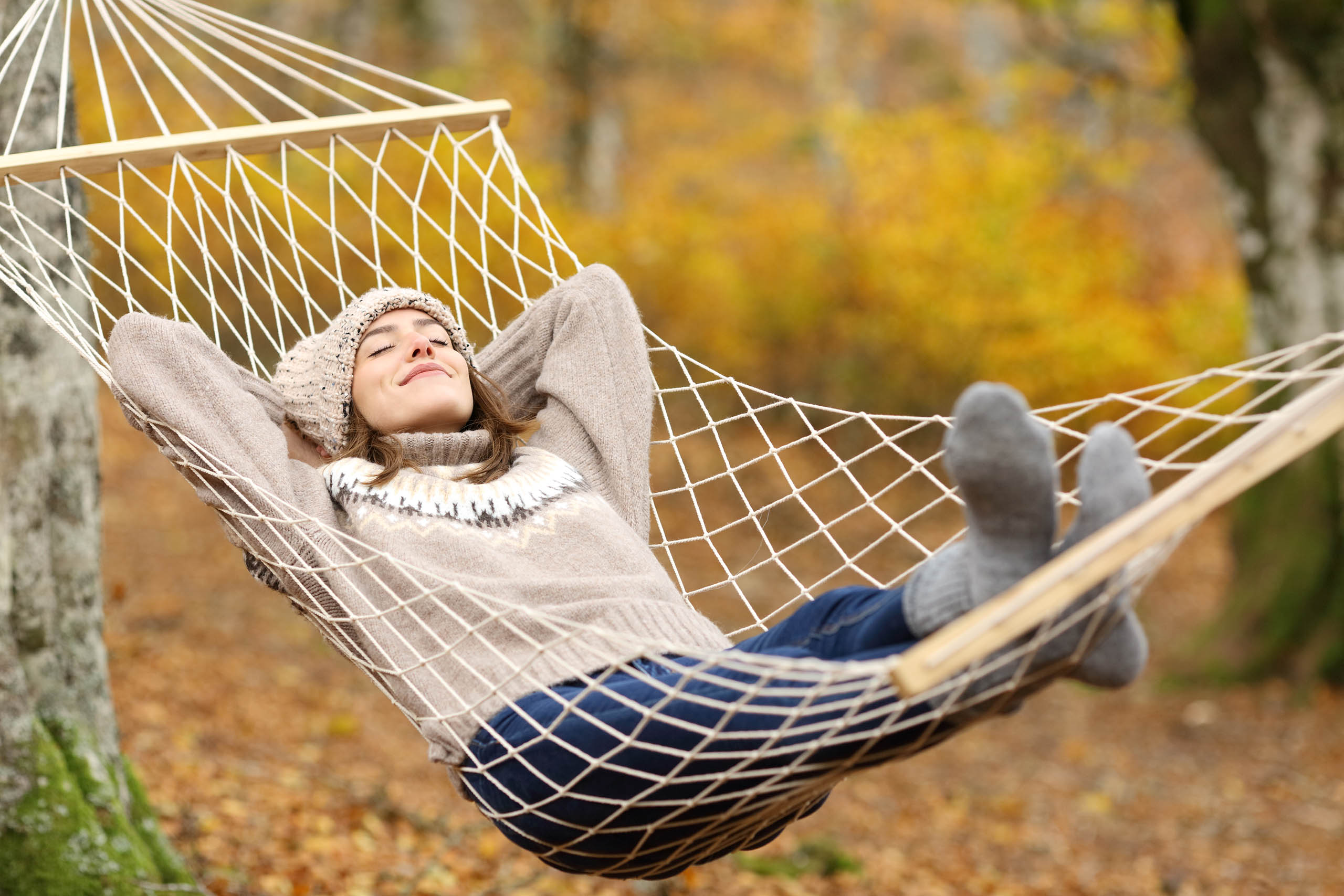 Young woman wearing a knit sweatshirt and hat lying happily in a net hammock outside during autumn.