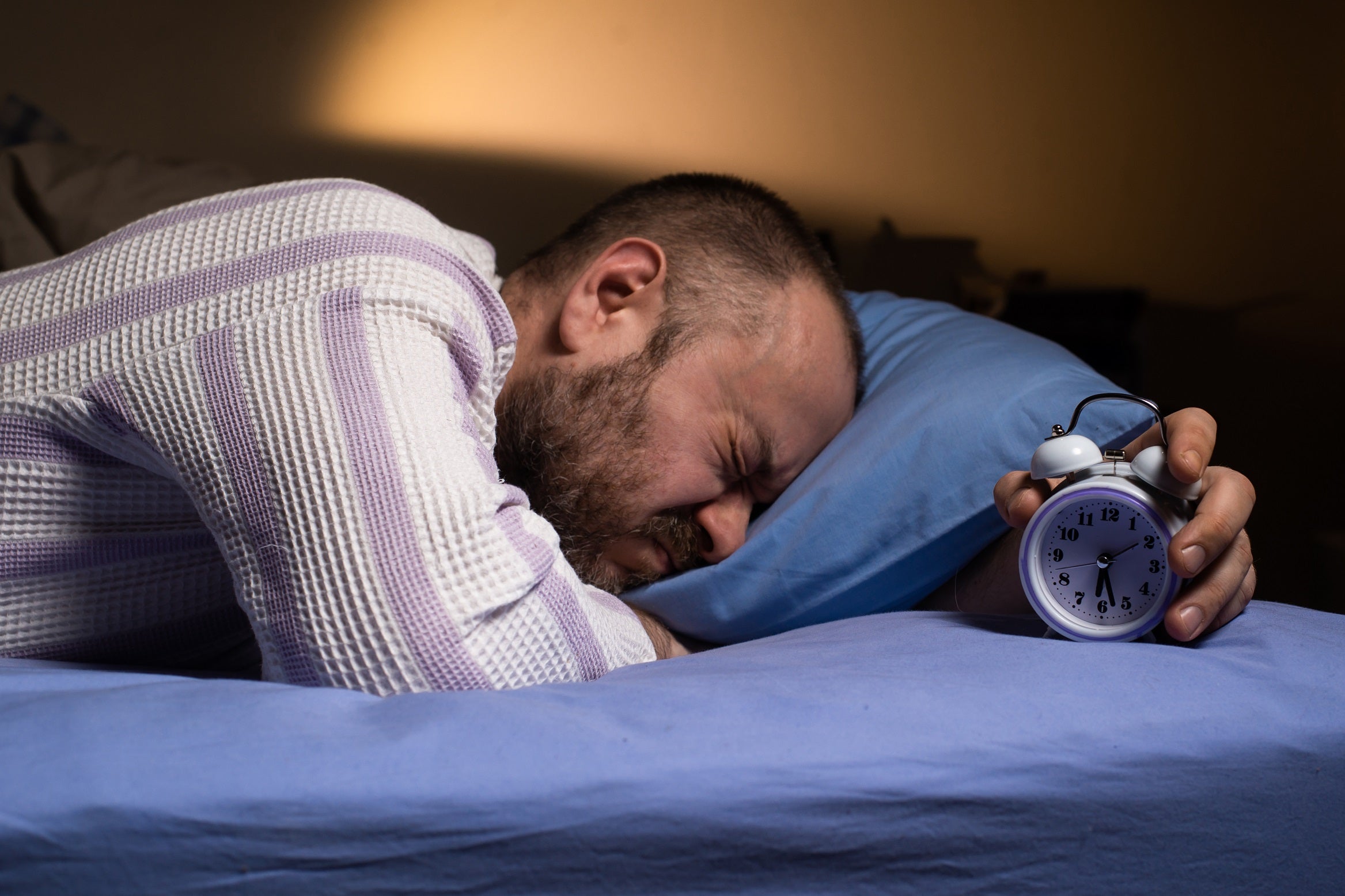 Man looking irritated while sleeping on blue sheets holding an alarm clock.