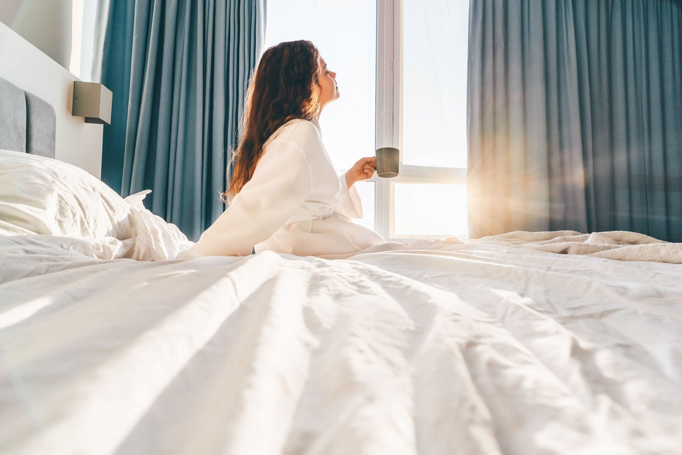 Femme assise dans son lit, tenant une tasse de café, le soleil entrant par la fenêtre.