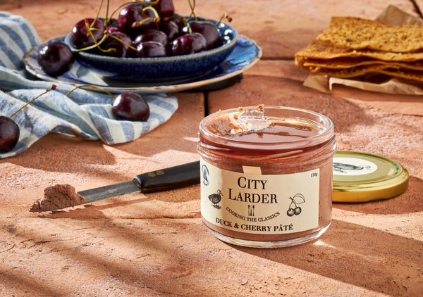 a jar of city larder duck and cherry pate in the sunlight accompanied by fresh cherries and a knife with some pate on it