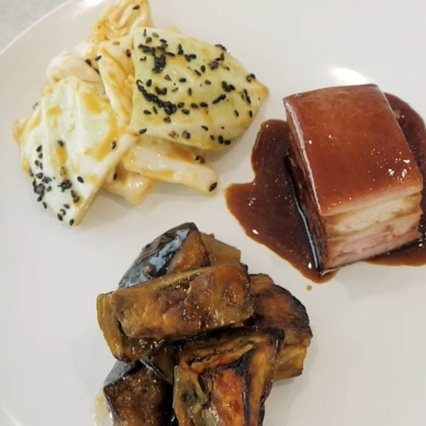 white plate with small amount of fodd white cabbage with black seeds a square of cooked pork in sauce and cubes of eggplant in sauce