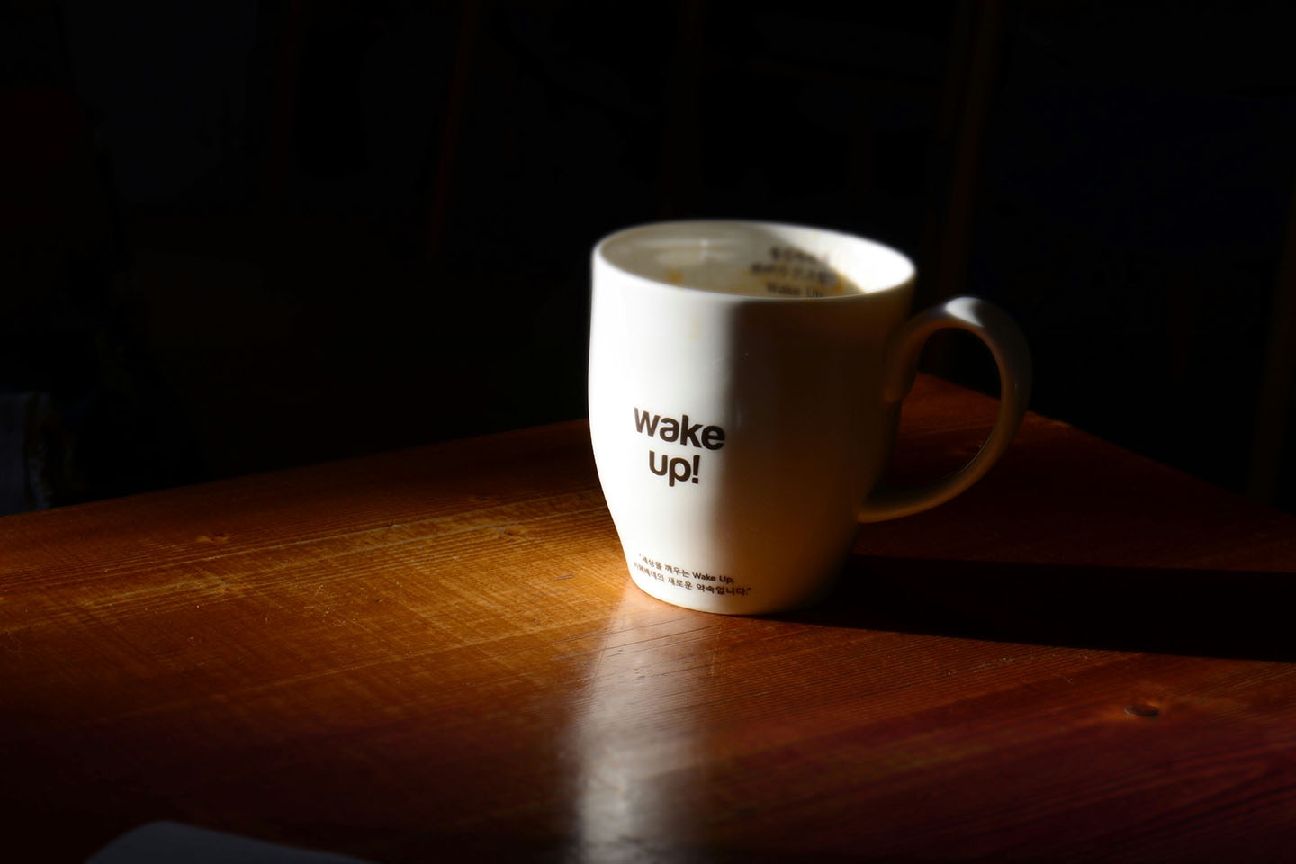 White coffee cup, against a black background on a wooden surface. Cup reads "wake up" in black letters on the front.