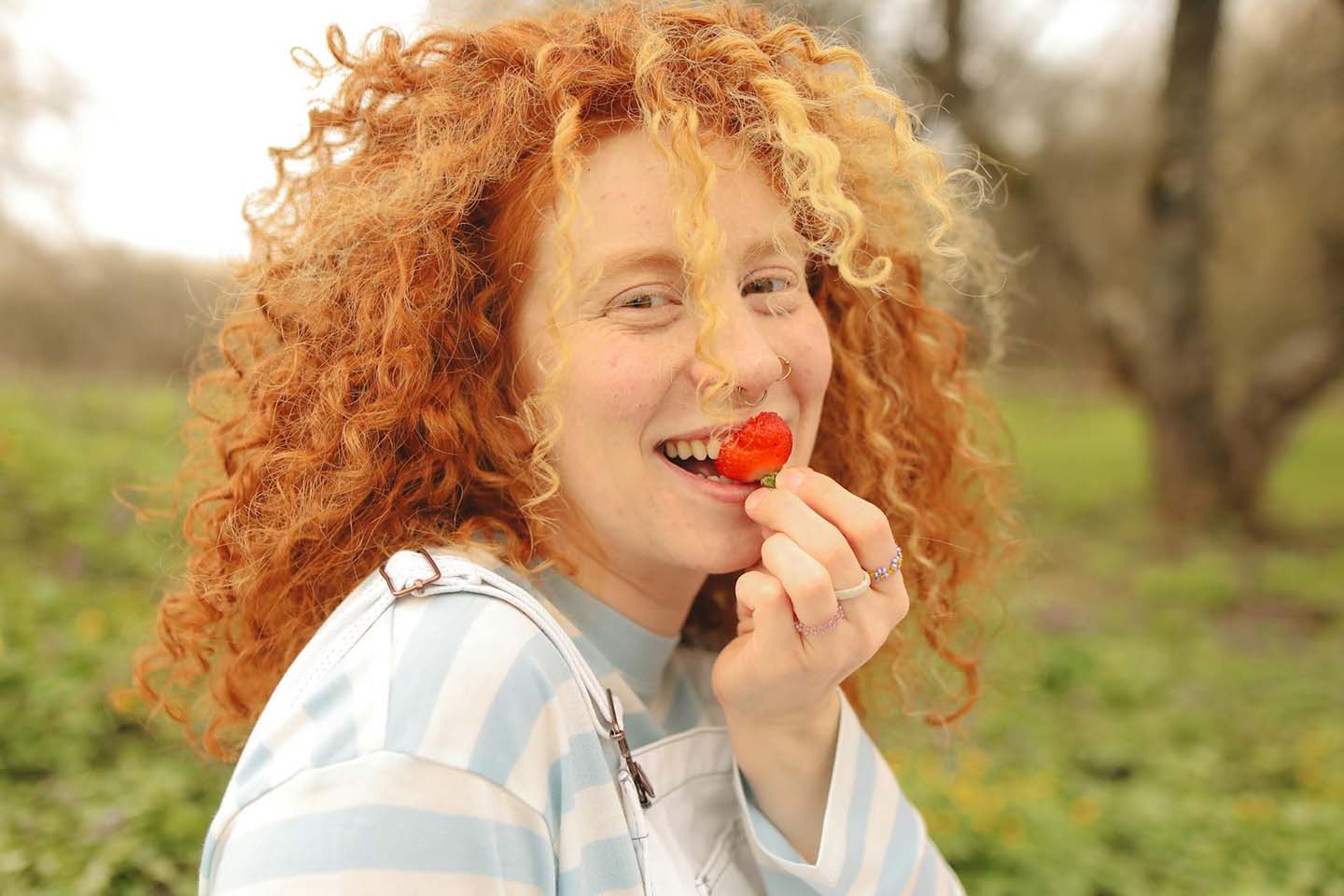 an adorable woman with curly red hair happily eating a strawberry