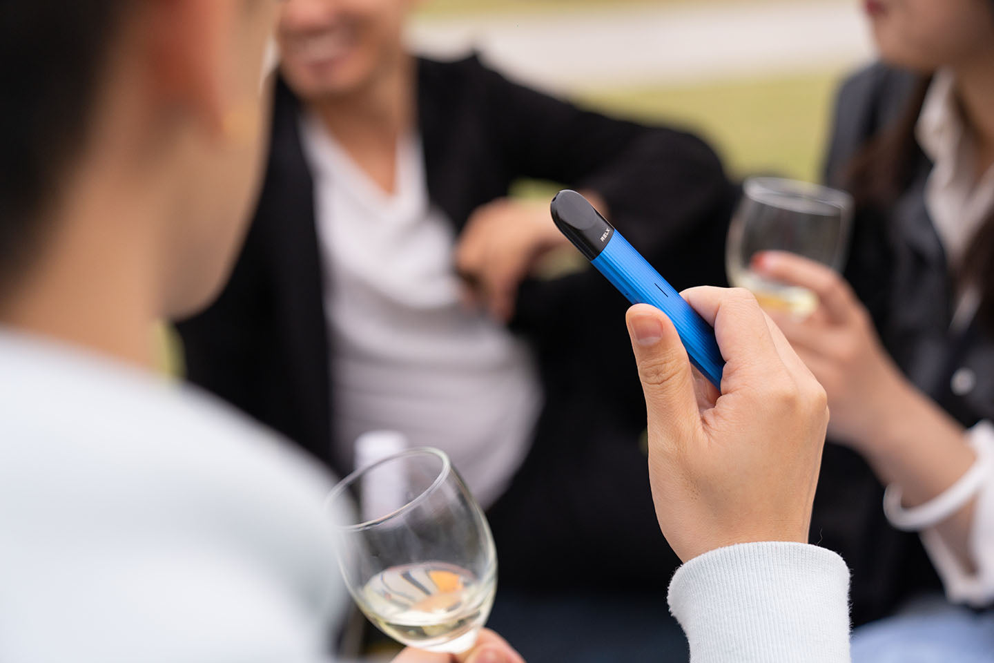 Three out of focus people sit drinking wine, while a hand holding a blue disposable vape is in focus in the foreground of the photo.