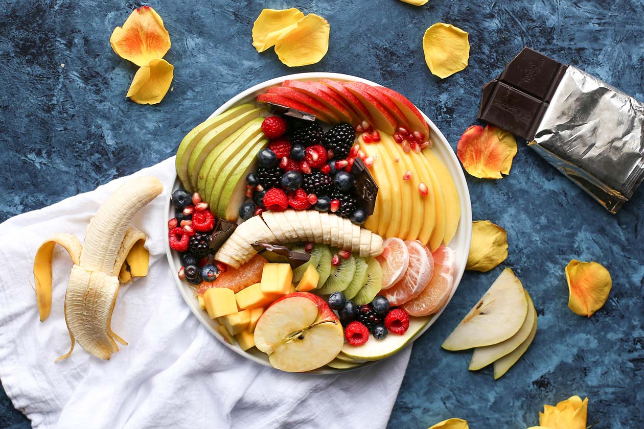 a bowl of assorted fruits beside a peeled banana and an opened chocolate bar on the table