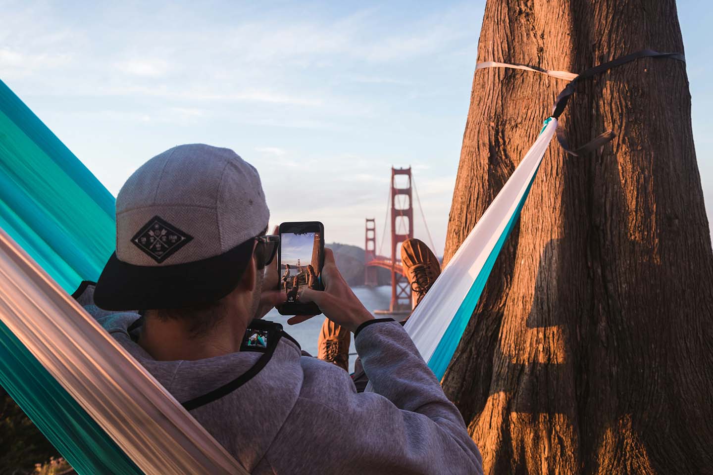 shallow focus photography of man on hammock while taking photo of golden gate bridge of california to his phone