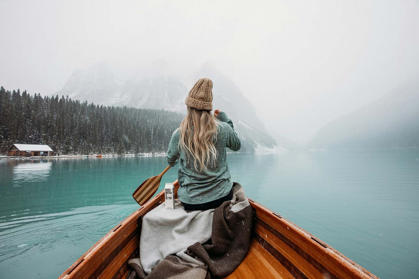 Woman sits at the front of a wooden boat rowing in the blue water with mountains and forests all around