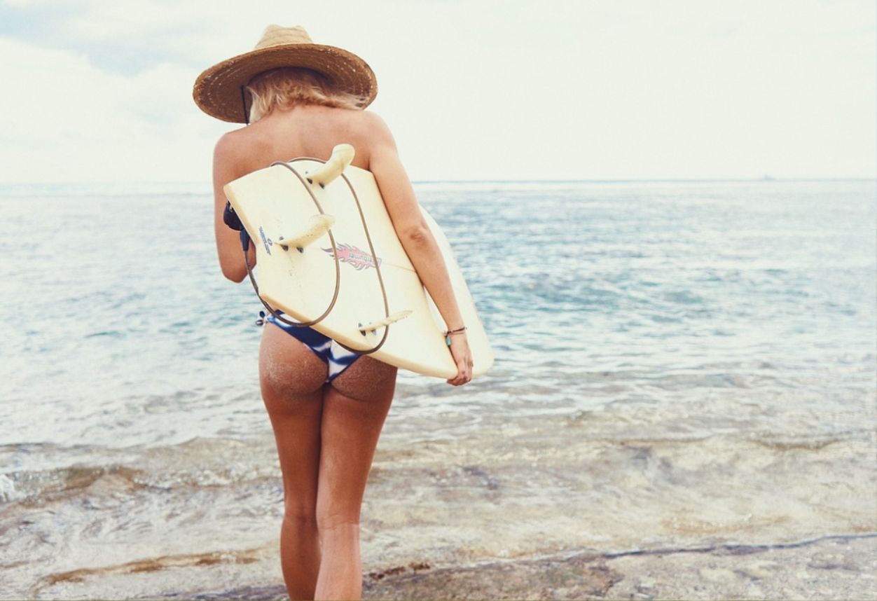 Girl in a bikini holding a surfboard and about to walk into the ocean with a big hat on.