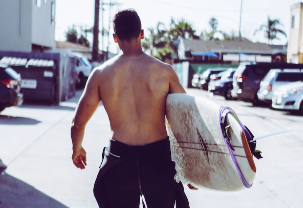 Guy with a half folded down wetsuit walking his foam surfboard down to the beach in San Diego.