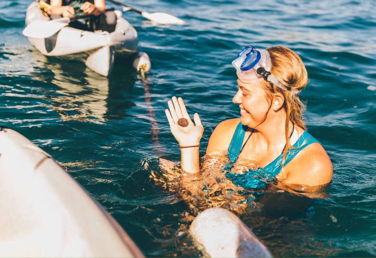 Chica rubia sosteniendo un dólar de arena vivo mientras está en el agua rodeada de kayaks