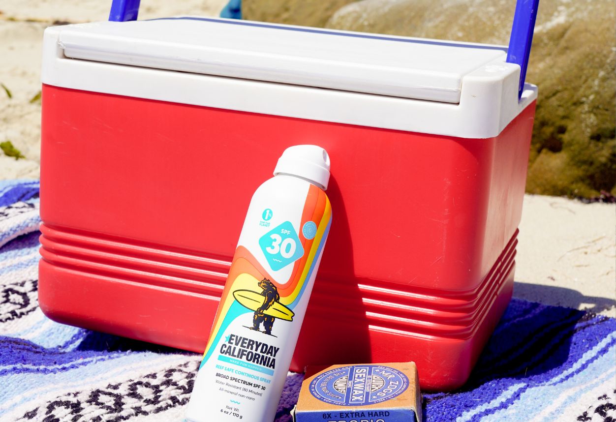 small red cooler sitting on the sand with a Baja blanket underneath with sunscreen and wax.