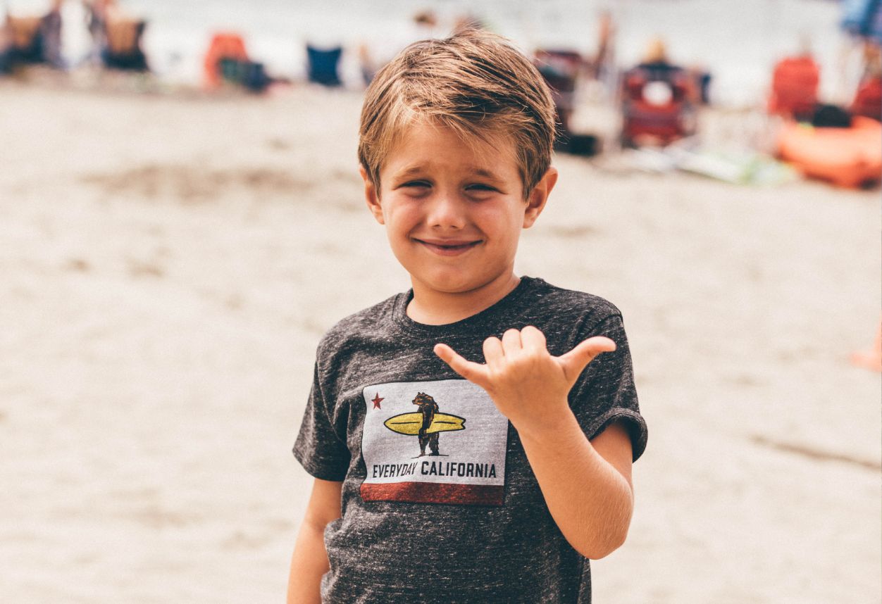 Niño en la playa vistiendo ropa cotidiana de California sosteniendo un Shaka